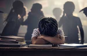 A student sits alone with his head down on his desk while shadowy classmates point and jeer around him